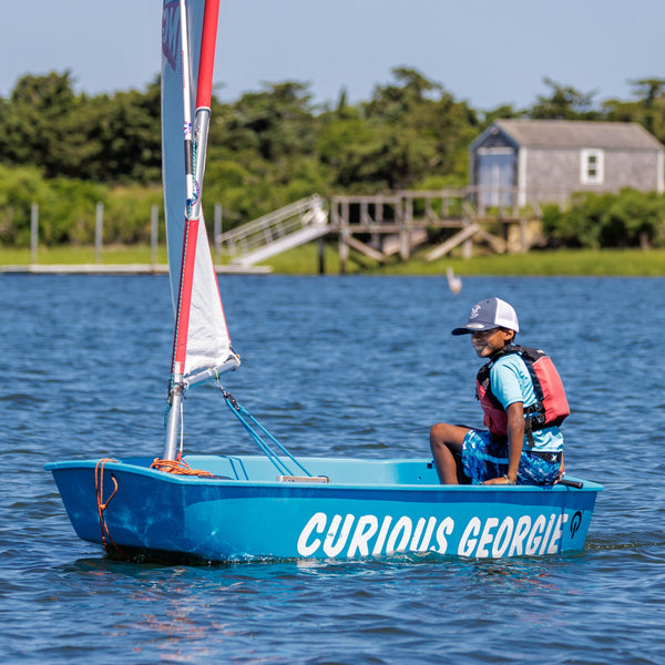 Child on a small sailboat named 'Curious George' on a body of water with trees and a building in the background.