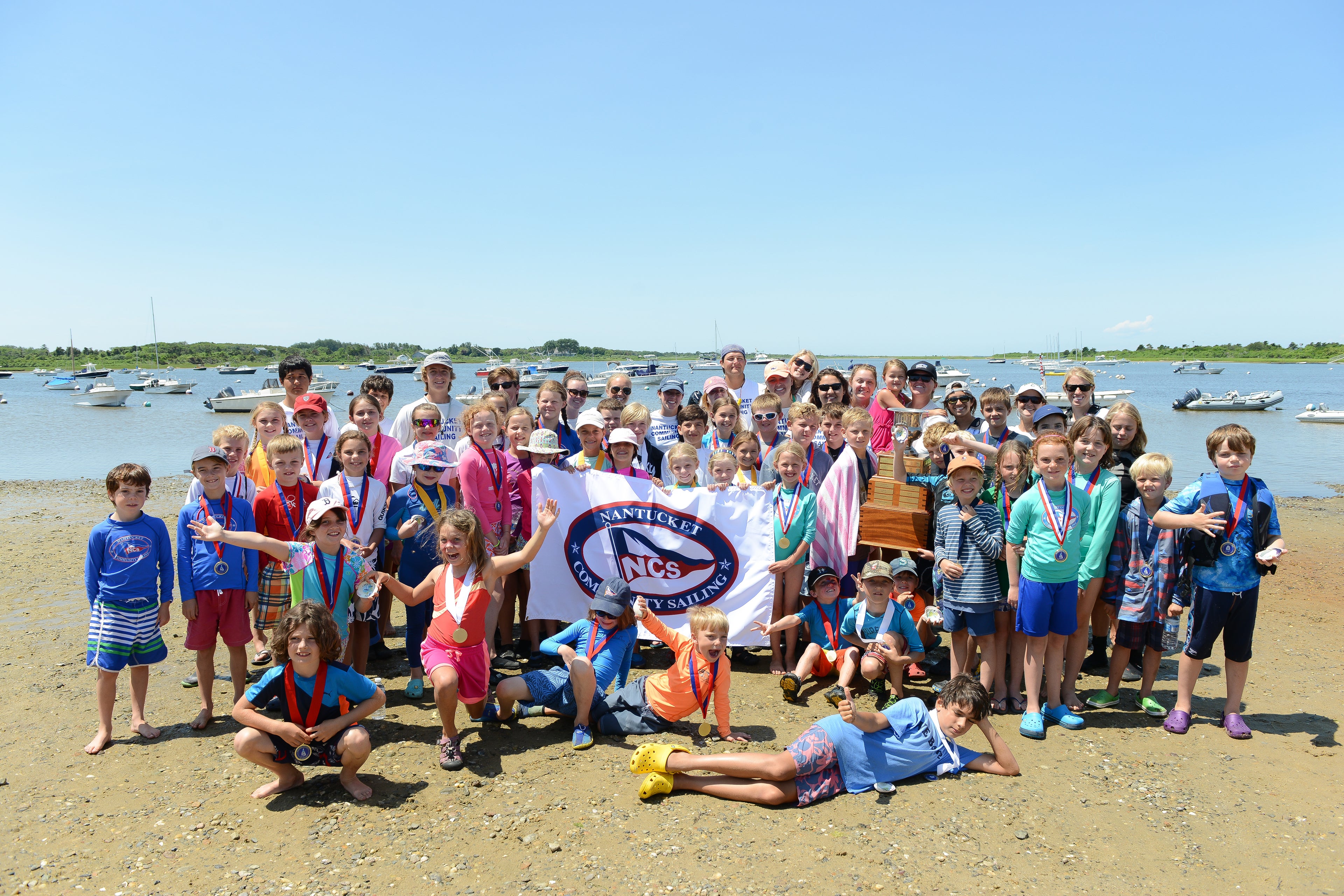 Group of children and adults standing on a beach holding a banner with 'Nantucket Community Sailing' on a clear day.