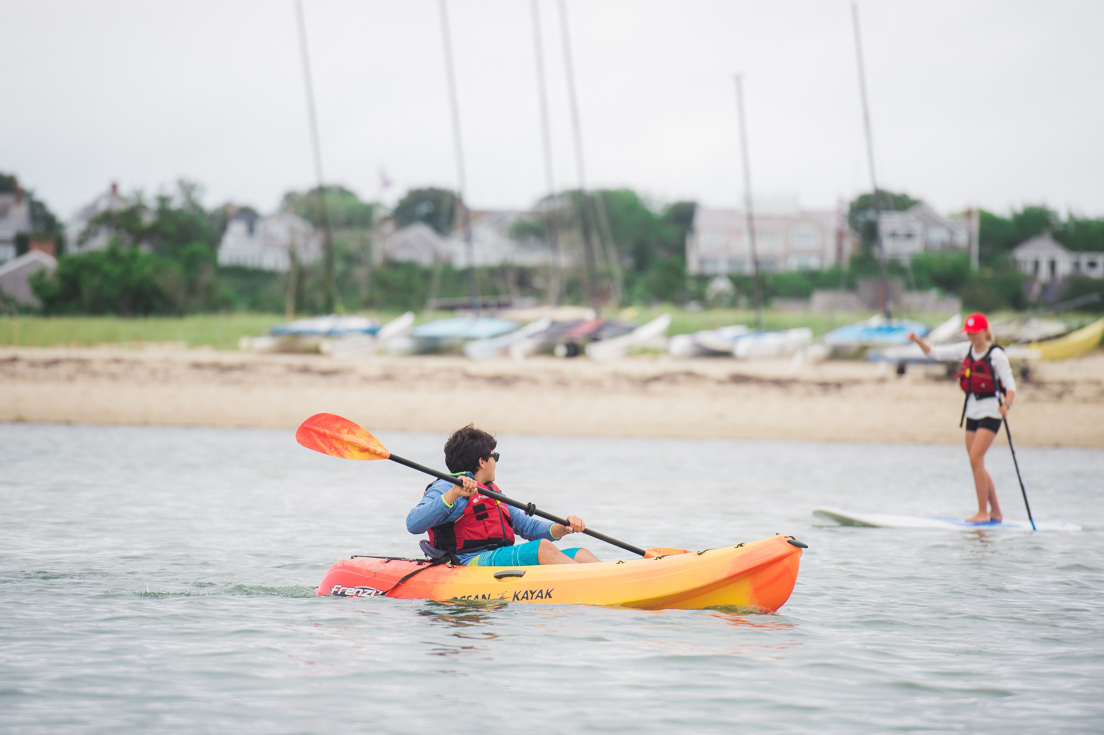 Two people on water, one in a kayak and the other on a stand-up paddleboard, with a beach and buildings in the background.