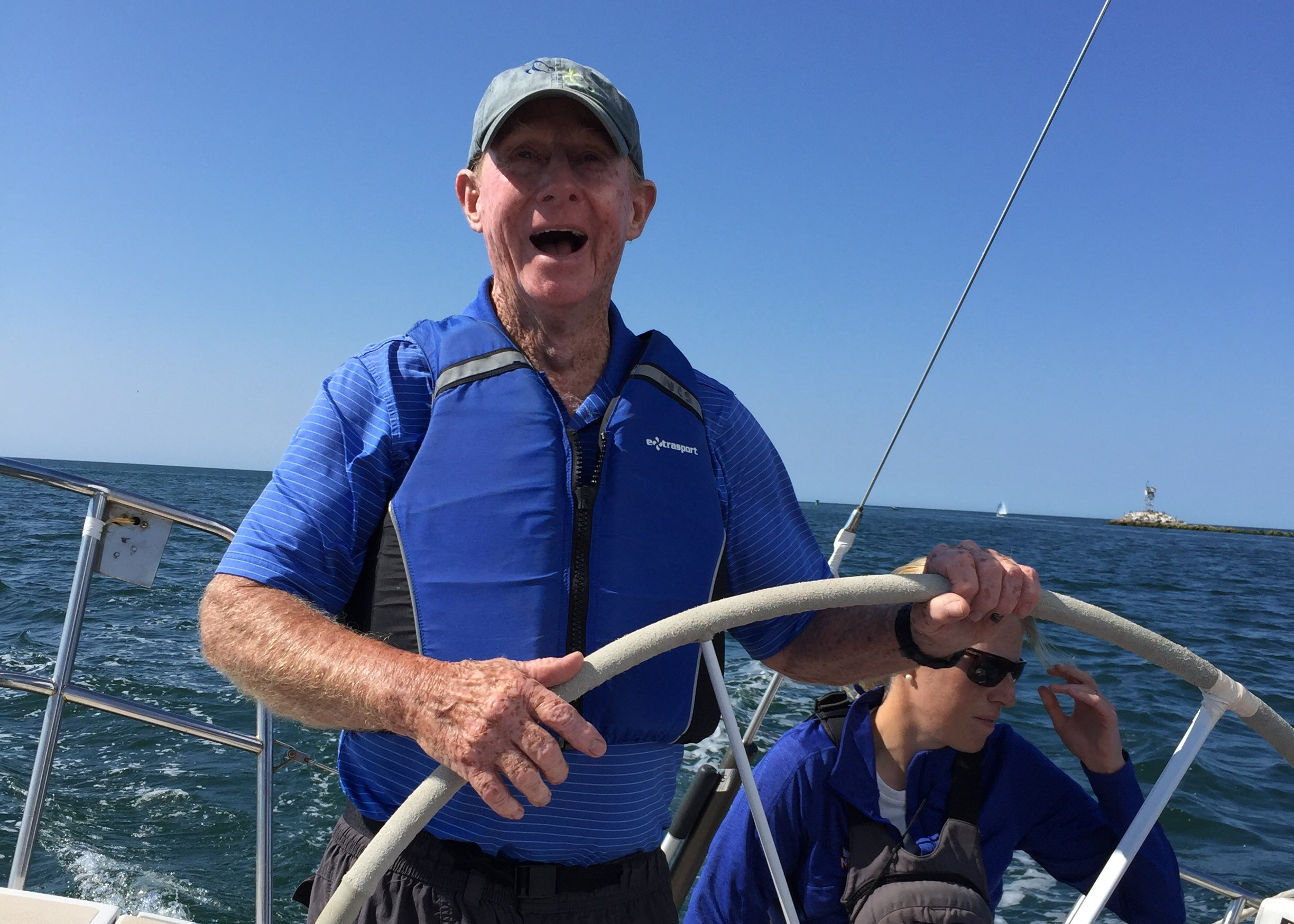 Man steering a sailboat with another person in the background on a clear day.