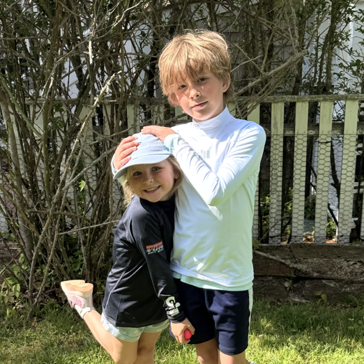 Two children playing outdoors in a grassy area with trees and a fence in the background.