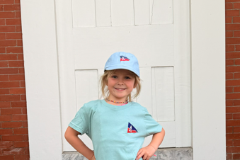 Young girl standing on steps wearing a light blue t-shirt with a logo and a matching cap.