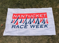 A white fabric flag or towel printed with 'NANTUCKET RACE WEEK' in blue and red, laid flat on green grass.