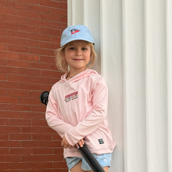 Child wearing a pink hoodie and blue cap leaning against a white wall.