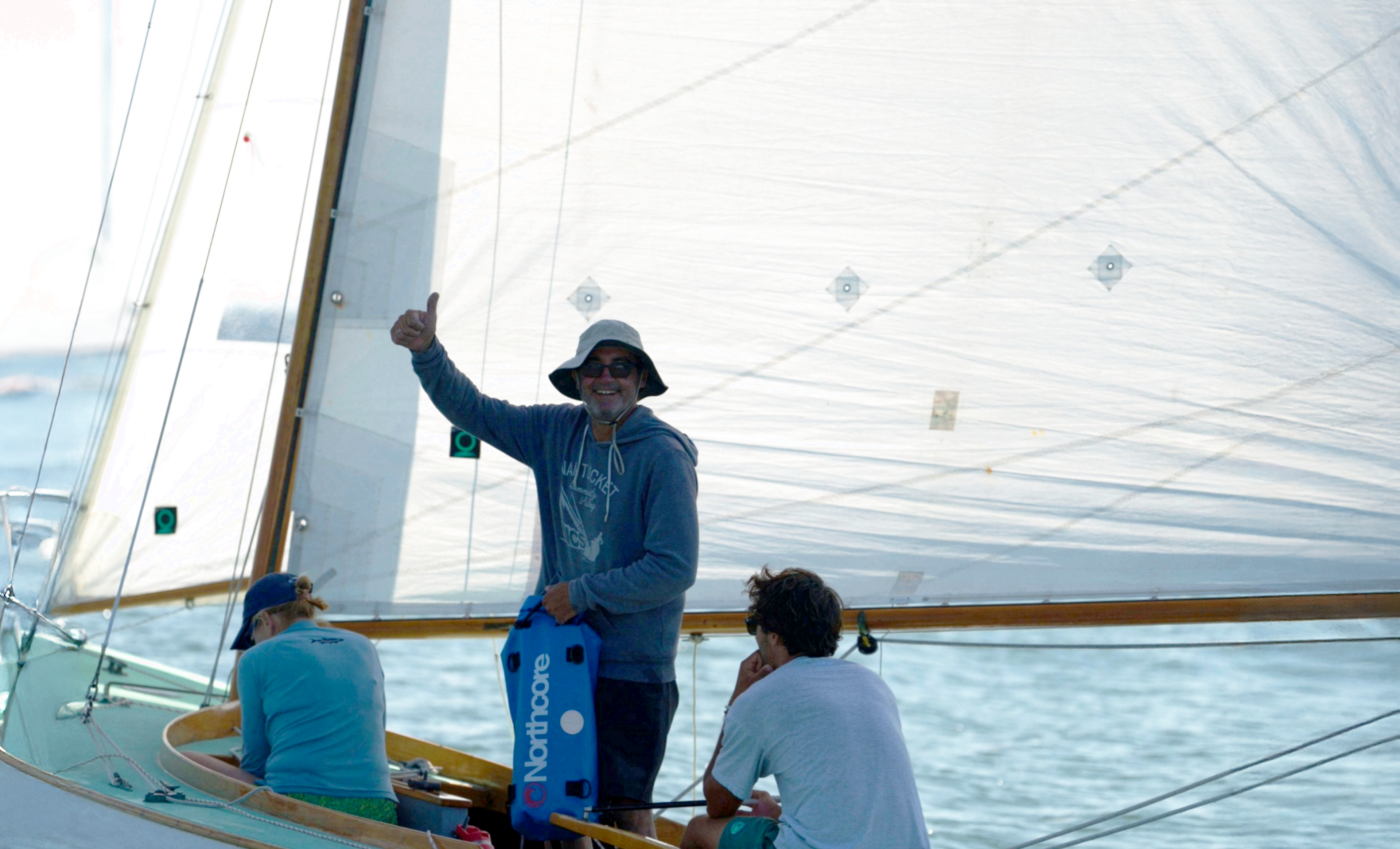 Person on a sailboat with a large white sail, smiling and holding onto the mast.