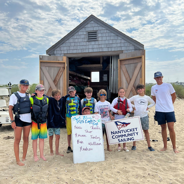 Group of children and adults standing in front of a wooden shed on a sandy beach, with signs and a Nantucket Community Sailing logo.